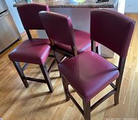 Three burgundy leather-like bar stools arranged on wooden floor near kitchen counter.