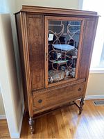 Full view of the wood china cabinet showing its shape, color, glass door with carved design, drawer below and turned legs.