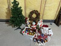 Wide view of Christmas decorations including the green artificial tree, wreath, assorted ornaments, and ceramic Santa Claus figurines on a concrete floor with unfinished wall background.