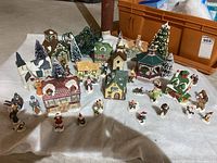 Full layout of Christmas village buildings, figurines, trees, and string lights spread on white cloth with orange tote container in background.