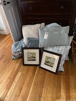 Full view showing two framed bathroom-themed art prints, six decorative pillows of various textures and colors including teal, blue, gray tones, fleece sheet set and throws, and knitted lap blanket piled on the floor.