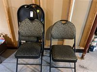 Four folding chairs, black metal frame with greenish-gray patterned upholstery on seats and backs, stacked and standing on concrete floor in unfinished basement.