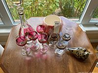Clear decanter, red-tinted floral glasses, clear snifter glasses with gold rim, spotted glass bird, and bowl on wooden table near window