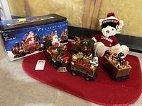 Four piece holiday train set displayed with original box and white plush bear wearing Canada sweater and earmuffs on red cloth.