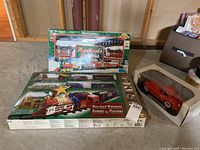 Photo showing two boxed holiday-themed model train sets and a boxed red Jaguar car model sitting on the floor in a basement setting.