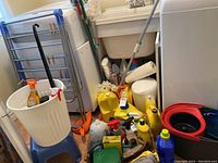 Overview of laundry room corner showing bucket on stool, cleaning supplies, mop heads, and yellow bottles.