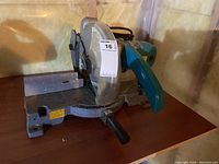 Front side view of Makita miter saw showing metal base, aqua blue handle, cutting blade and adjustment handle