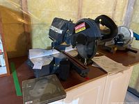 Angle view of large black and metal mitre saw with Craftsman dust collection bag, sitting on cabinet.
