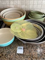 Multiple mixing bowls grouped: ceramic nested bowls with colored rims, green plastic bowls nested, metal bowls nested with yellow translucent leaf-shaped plastic lids on top.