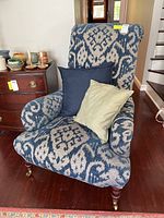 Full front view of the upholstered armchair showing ikat pattern, rolled arms, wood legs, and included pillows