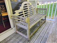 Front and side angle view of used teak patio bench on wooden deck. Weathered grey wooden slats and curved decorative backrest visible.