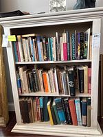 Photo of a white bookshelf containing three shelves full of assorted books showing wear and tear.