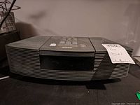 Front view of Bose Wave Radio/CD player, showing horizontal speaker vents and control panel buttons on top, dusty surface.