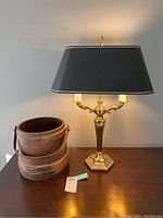 Wooden bucket next to brass finish lamp with black shade on wooden table.