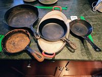 Five cast iron cookware pieces arranged on counter. Large and medium round skillets plus small skillets and rectangular griddle with chipped enamel handles visible. Rust and scratch wear visible.