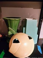 Three pottery vases on black shelf: green vase with fish motif, blue geometric vase, beige round vase with cut-outs.