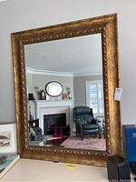 Large beveled edge mirror with ornate gold frame shown hanging on a wall reflecting a living room with fireplace.
