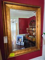 Framed wall mirror mounted on a red wall reflecting part of an interior room including furniture and framed artwork on the floor.