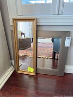 Two mirrors leaning on a wall, one with a gold detailed frame and the other with a plain grey frame.