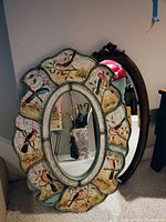 Two oval framed mirrors leaning against a wall; one painted with birds and foliage and one carved wood frame with ornamental detail.