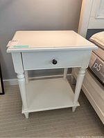 Front view of a white night stand with drawer and lower shelf, showing turned legs and black knob drawer handle.