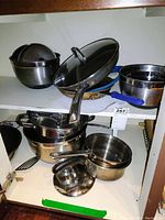 Shelf containing various stainless steel pots, pans and bowls, some with glass lids, some with blue handles. Items show scratches and visible wear.