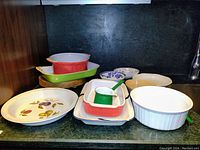 Photo showing various baking dishes including a large oval with fruit design, several rectangular and round dishes, two blue and white small patterned bowls, enamelware ladle, and wood cutting board.