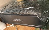 Side view of black electric smokeless grill covered in plastic wrap, showing 'POWER SMOKELESS GRILL' branding on the front.