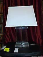 Front view of glass table lamp showing rectangular clear glass base and off-white rectangular shade on black surface with burgundy curtain background.