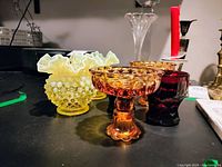 Yellow hobnail style glass vase with ruffled edge, two translucent amber glass compotes with pedestal bases, and a deep red ruby glass tumbler on a dark surface.
