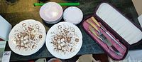 Photo showing two stacks of ceramic plates, one larger with brown floral pattern, one smaller with various smaller plates, and a three-piece carving set in a worn maroon case.