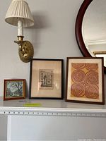 Three framed wall art pieces placed on mantel under a wall sconce and mirror. Includes cityscape, architectural print, and spiral design.