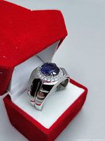 Front angled view of men's silver ring showing large oval blue sapphire with clear stone halo, polished chunky band, red velvet ring box interior.