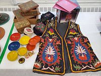 Photo of embroidered textile vest with colorful floral embroidery, assorted hand-painted keepsake boxes, lacquerware bowls, and miniature nested dolls on display.