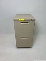 Front view of beige metal 2 drawer file cabinet with yellow lot sticker on top drawer, showing recessed handles and keyhole.