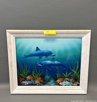 Framed oil painting showing two dolphins underwater with aquatic plants and coral, in a whitewashed wooden frame.