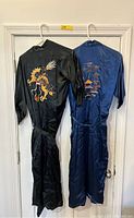 Full view of two silk Chinese kimono robes hung on a wall. One black with dragon embroidery, one blue with a Chinese landscape embroidered scene.
