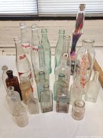 Full view of assorted vintage beverage, medicine, and apothecary bottles on a white surface with window blinds background