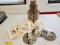 Full view of the Sunbeam food processor base unit with brown plastic bowl and handle, three stainless steel discs arranged beside it, white food pusher, and instruction books laid out on table.