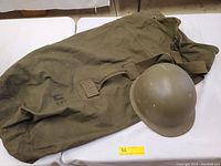 Canvas duffel bag and army helmet together on a white surface. Bag appears olive green with a US marking and leather/canvas straps. Helmet is olive drab with visible wear.