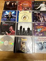Photo showing 12 CD cases arranged in 3 rows. Visible CDs: The Priests Vol 1, Traditional Irish Dance Music, Andre Rieu Greatest Hits, Boney M, Classics at the Pipes, La Boheme, Classic Rock 70s, The Priests black and white photo, Classic Rock compilation, Joseph musical soundtrack, Christmas Legends, and another Priests album.