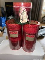 Photo showing three candles: one large glass container candle in red and green with holiday tag, and two smaller red pillar candles labeled 'Holiday Magic'