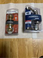Two door locks side by side on a wooden floor: left is a polished brass keyed entry knob by Gate House in original packaging; right is a chrome lever-style passage lock by Elements Weiser in original packaging.
