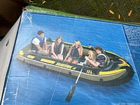 Box image showing the boat with 4 people on a lake, illustrating size and design, with two oars attached.
