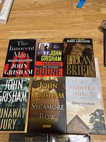 Eight John Grisham hardcover books displayed on a wooden surface showing front covers including The Innocent Man, Theodore Boone, The Pelican Brief, The Runaway Jury, Sycamore Row, A Painted House, The Associate, and The Racketeer.