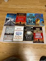 Photo showing 9 softcover John Grisham books arranged on wooden floor, titles visible.