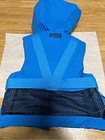 Back view of the blue kids life jacket showing Y-shaped blue webbing straps, hood, and mesh lower back panel.