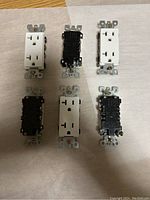 Top view of six electrical outlets: three outlets with white faceplates and three with black faceplates arranged on a flat surface.