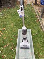 Full view of the white Tineco hard floor cleaner standing upright on a plastic container outdoors on grass with leaves.