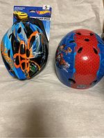 Two children's helmets side by side on beige fabric background, left is Hot Wheels blue and black with orange accents, right is Paw Patrol blue with red stripe and character graphics.